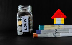 A lego home on top of 3 money bundles and a glass jar full of coins labelled as loan signifying whether can I take home loan after personal loan.